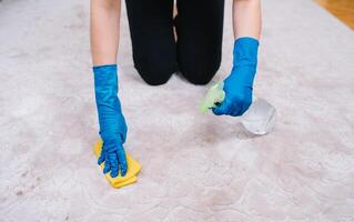 personnes, travaux ménagers et Entretien ménager concept - proche en haut de femme dans caoutchouc gants avec tissu et détergent vaporisateur nettoyage tapis à maison. proche en haut de femme avec tissu nettoyage tapis photo