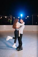 couple sur le ville patinoire dans une hiver soir. gars portion agréable fille à patin sur le la glace dans le foncé nuit et scintille éclairage au dessus leur photo