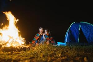 nuit camping dans le montagnes. content couple voyageurs séance ensemble à côté de feu de camp et embrasé touristique tente. sur Contexte gros rocher, forêt et nuit ciel. photo
