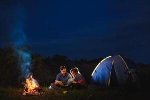 Jeune couple homme et femme voyageurs séance près embrasé touristique tente, brûlant feu de camp, sur le Haut de montagne, profiter magnifique vue de nuit ciel. photo