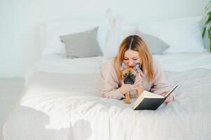 une femme en train de lire une livre et souriant comme elle est assis dans lit. le alarme l'horloge sur le bureau à côté de son. photo