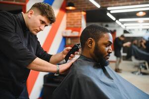 portrait de Jeune noir homme étant taillé avec professionnel électrique tondeuse machine dans salon de coiffure.male beauté traitement concept. Jeune africain gars avoir Nouveau la Coupe de cheveux dans coiffeur salon photo