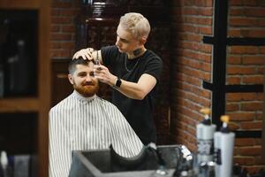 fabrication la Coupe de cheveux Regardez parfait. Jeune barbu homme avoir la Coupe de cheveux par coiffeur tandis que séance dans chaise à salon de coiffure. photo