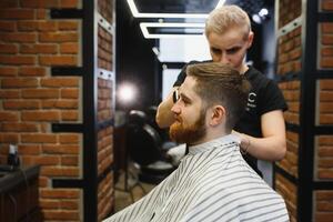 fabrication la Coupe de cheveux Regardez parfait. Jeune barbu homme avoir la Coupe de cheveux par coiffeur tandis que séance dans chaise à salon de coiffure. photo