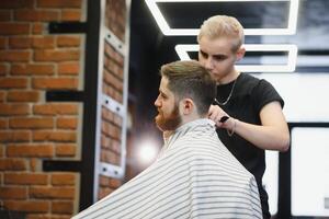 fabrication la Coupe de cheveux Regardez parfait. Jeune barbu homme avoir la Coupe de cheveux par coiffeur tandis que séance dans chaise à salon de coiffure. photo