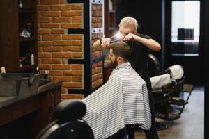 fabrication la Coupe de cheveux Regardez parfait. Jeune barbu homme avoir la Coupe de cheveux par coiffeur tandis que séance dans chaise à salon de coiffure. photo