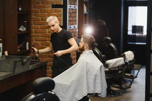 fabrication la Coupe de cheveux Regardez parfait. Jeune barbu homme avoir la Coupe de cheveux par coiffeur tandis que séance dans chaise à salon de coiffure. photo