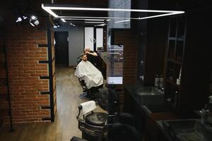 fabrication la Coupe de cheveux Regardez parfait. Jeune barbu homme avoir la Coupe de cheveux par coiffeur tandis que séance dans chaise à salon de coiffure. photo