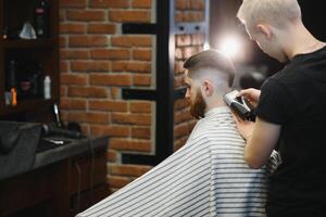 Jeune Beau coiffeur fabrication la Coupe de cheveux de attrayant barbu homme dans salon de coiffure. photo