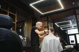 fabrication la Coupe de cheveux Regardez parfait. Jeune barbu homme avoir la Coupe de cheveux par coiffeur tandis que séance dans chaise à salon de coiffure. photo