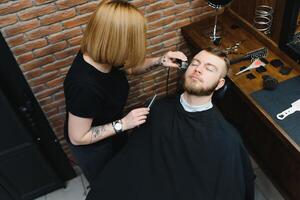 qualifié femelle coiffeur Coupe une barbe de brunette adulte gars dans professionnel rétro salon de coiffure. photo