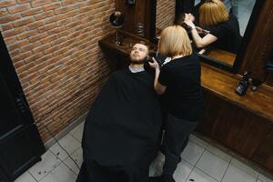 qualifié femelle coiffeur Coupe une barbe de brunette adulte gars dans professionnel rétro salon de coiffure. photo