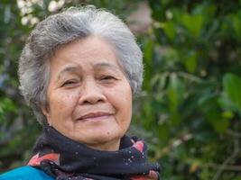 portrait de une content Sénior femme souriant et à la recherche à le caméra tandis que permanent contre des arbres. concept de vieilli gens et soins de santé photo