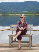 portrait de Sénior asiatique femme portant des lunettes de soleil et à la recherche à le caméra tandis que séance sur une en bois chaise côté le étang. concept de vieilli gens et relaxation photo