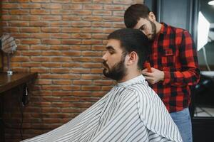 sérieux barbu homme avoir barbe la Coupe de cheveux avec une tout droit le rasoir par coiffeur tandis que séance dans chaise à salon de coiffure. salon de coiffure thème. photo