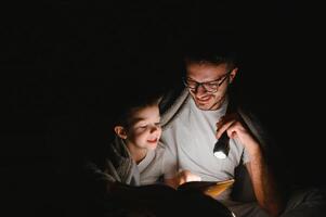 content famille en train de lire heure du coucher récit en dessous de couverture dans soir. père et fils dépenser temps ensemble. du père journée photo