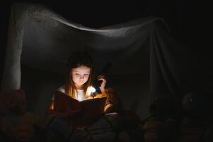 peu fille sont en train de lire une livre avec lampes de poche dans tente. content fille en jouant à maison. marrant charmant enfant ayant amusement dans chambre. photo