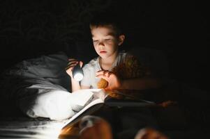 enfant en train de lire livre dans lit. des gamins lis à nuit. peu garçon avec Fée conte livres dans chambre . éducation pour Jeune les enfants. heure du coucher récit dans le soir. mignonne enfant en dessous de couverture dans foncé pièce avec lampe photo