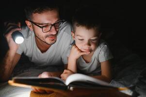 père et le sien peu fils en train de lire heure du coucher récit à maison. photo