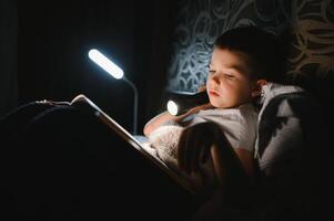 portrait de mignonne peu garçon en train de lire dans lit avec lampe de poche dans foncé chambre, profiter contes de fées photo