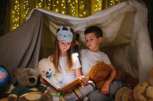 deux les enfants avec lampe de poche lis une livre en dessous de une couverture comme une tente photo