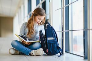 peu jolie fille séance sur le sol de le école couloir et en train de lire une livre. le concept de scolarité. photo