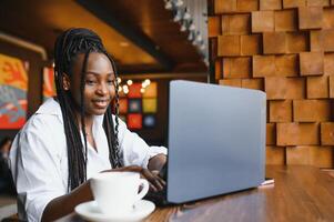 Jeune africain américain fille séance dans restaurant et dactylographie sur sa portable. jolie fille travail sur ordinateur à café. photo