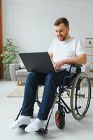 portrait de souriant désactivée Masculin séance dans fauteuil roulant et travail sur portable de maison. Jeune ouvrier avec spécial besoins. pigiste et gens avec handicapées concept photo