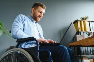 Jeune homme avec spécial Besoins dans décontractée vêtements travail sur sans fil portable. Masculin pigiste travail de Accueil tandis que séance dans fauteuil roulant photo