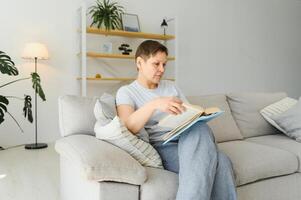 attrayant âge moyen femme avec une charmant sourire séance sur une canapé dans le vivant pièce agrippant une livre. photo