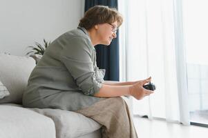 une très émotif plus âgée Dame est assis sur le canapé à Accueil et pièces Jeux. grand-mère gagne tandis que séance seul à maison. photo