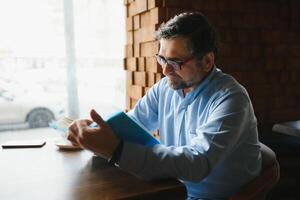 Sénior vieux homme en train de lire une livre dans une café boutique, profiter le sien littéraire loisir photo