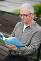 vieux homme avec gris cheveux lit une livre sur une banc dans le parc. du repos dans le parc. photo