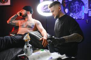 barbu tatouage artiste travail à le sien studio tatouage manche sur le bras de le sien Masculin client. homme avoir tatoué par professionnel tatoueur photo