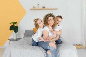 portrait de souriant Jeune maman embrassement des gamins peu fils et fille. content famille. mère avec les enfants séance sur lit et aimant le bien fois ensemble. fermer photo