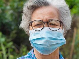 portrait de un personnes âgées femme portant une visage masque à la recherche à le caméra tandis que permanent dans une jardin. masque pour protéger virus, corona virus, pollen céréales. concept de vieux gens et soins de santé photo