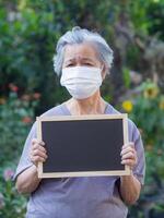 portrait de un personnes âgées femme en portant une tableau noir et portant une visage masque pour protéger virus, COVID-19 [feminine, corona virus, bactéries, pollen céréales, air la pollution pm 2.5, et plus. soins de santé concept photo