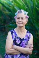 portrait de une Sénior femme cheveux bigoudi, bras franchi, souriant, et à la recherche à le caméra tandis que permanent dans une jardin. espace pour texte. concepts de vieilli gens et soins de santé photo