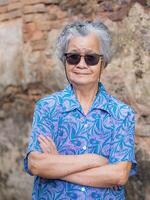 Sénior femme avec court gris cheveux portant des lunettes de soleil, bras franchi, souriant, et à la recherche à le caméra tandis que permanent avec vieux brique mur Contexte. concept de vieilli gens et relaxation photo