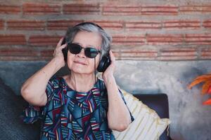 un personnes âgées femme avec court gris cheveux, portant sans fil écouteurs écoute à une préféré chanson tandis que séance dans le chambre. espace pour texte. concept de vieilli gens et relaxation photo