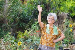 portrait de un personnes âgées femme des exercices par faire des gestes élever tous les deux bras, souriant, et à la recherche à le caméra tandis que permanent dans une jardin. espace pour texte. concept de vieilli gens et soins de santé photo