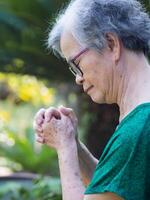 une portrait de un personnes âgées femme mains rejoint ensemble tandis que permanent dans une jardin photo