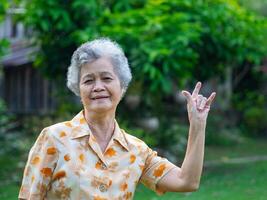 portrait de une content Sénior femme montrant mains l'amour vous symbole tandis que permanent dans le jardin. espace pour texte. concept de vieilli gens et soins de santé photo