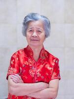 un personnes âgées femme portant une traditionnel cheongsam qipao robe, souriant et bras franchi tandis que permanent à maison. chinois femme dans rouge costume Nouveau année fête. concept de vieilli gens chinois photo