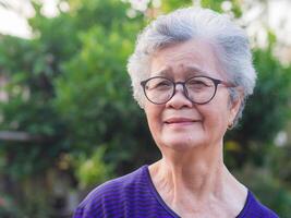 fermer de visage personnes âgées femme souriant bonheur, court blanc cheveux et à la recherche à le caméra, permanent dans jardin. asiatique Sénior femme en bonne santé et avoir positif pensées sur la vie faire sa content chaque journée photo