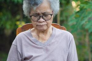 personnes âgées femme avec court blanc cheveux, portant lunettes, œil fermé et séance sur chaise dans le jardin. asiatique Sénior femme inquiet à propos sa santé problèmes. santé se soucier concept photo