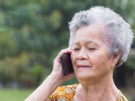 Sénior femme parlant sur sa mobile téléphone souriant avec délice comme elle écoute à le conversation tandis que permanent dans une jardin. espace pour texte. concept de vieilli gens et la communication photo