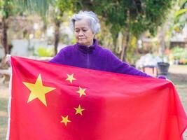 portrait de un personnes âgées asiatique femme avec court gris cheveux en portant une chinois drapeau tandis que permanent dans une jardin. concept de vieilli gens et relaxation photo