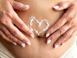 proche en haut de les femmes mains milieu âge avec blanc ongles appliquant crème dans le forme de cœur sur sa estomac. soins de la peau, soi se soucier, et personnel hygiène avec sur l'amour et corps positivité. photo