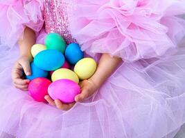 enfant dans rose tulle robe détient dans sa mains poignée de brillant multi coloré Pâques œufs, symbolisant le printemps vacances et le fête de Pâques. photo